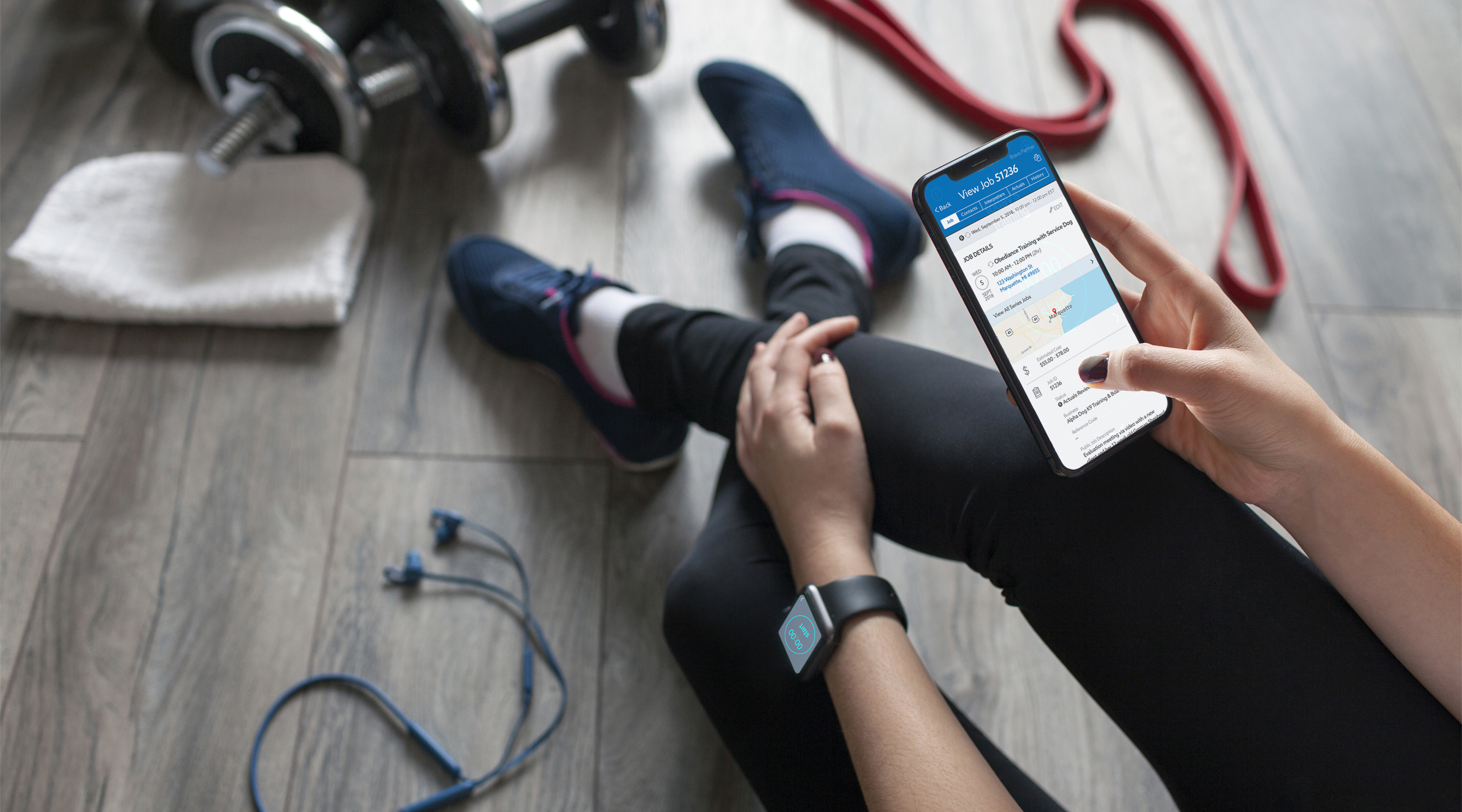 Woman sitting on the floor of a gym, presumably taking a rest after working out. She has her legs casually crossed and is surounded by workout equipment. She is holding her iPhone in her right hand with the Terp Scheduler iOS app for Agencies open viewing job details.